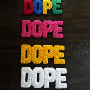 May include: Four embroidered patches featuring the word DOPE in block letters. The top patch has multicolored letters in red, blue, yellow, and green. The second is solid pink, the third is solid yellow, and the bottom patch is solid white.