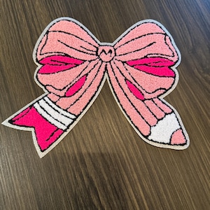 May include: A pink bow-shaped rug with a textured surface. The bow features a lighter pink center and darker pink accents, outlined in black. The bow's ends are designed to resemble a pencil, with white and pink stripes.