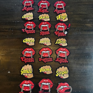 May include: A collection of embroidered patches featuring red lips with gold chain accents. Each patch displays a zodiac sign and the word "queen" in red script. The patches are arranged on a dark wooden surface.