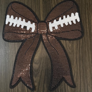 Puede incluir: Un lazo marrón con lentejuelas y costuras blancas, que se asemeja a un balón de fútbol americano. El lazo tiene un contorno negro y está sobre un fondo de madera. Ideal para manualidades y accesorios con temática deportiva.