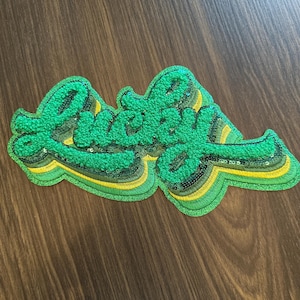 May include: A green embroidered patch with the word "Lucky" in a cursive font. The patch has a layered design with yellow and black accents and is embellished with sequins. The patch is on a brown wooden surface.