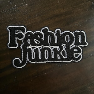 May include: Black and white embroidered patch with the words "Fashion Junkie". The patch has a black, fuzzy texture with a white border. The patch is on a dark brown wooden surface.