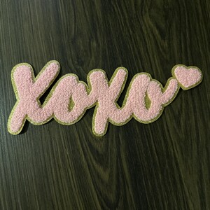 May include: Pink XOXO patch with a gold glitter border and a small heart. The letters are in a cursive style and have a textured, fuzzy appearance. The patch is likely for clothing or accessories.