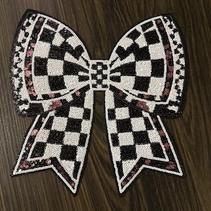 May include: A large, decorative bow applique with a black and white checkered pattern. The bow is outlined with black sequins and accented with pink sequins. The bow is likely for clothing or accessories.