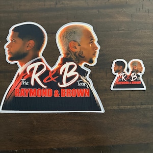 May include: Two die-cut stickers featuring profiles of two men, with the text The R&B Tour and RAYMOND & BROWN in red and white. The larger sticker is on the left and the smaller one is on the right, both showing the same graphic design.