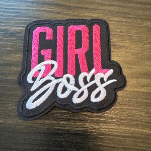 May include: Embroidered patch with the words "GIRL BOSS" in pink and white stitching on a black background. The patch has a black border and is likely designed to be ironed or sewn onto clothing or accessories.