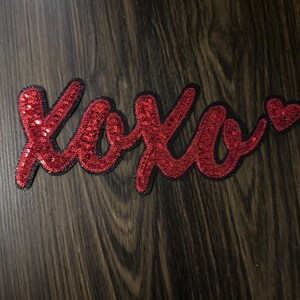 May include: Red sequined "XOXO" patch with a small heart. The letters are outlined in black and are on a dark wood grain surface. The patch is a decorative item.