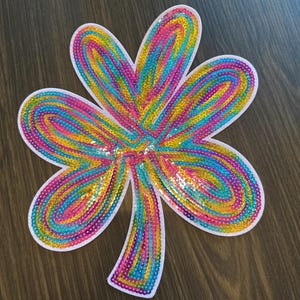 May include: A large, colorful sequined shamrock with a white border. The shamrock is filled with rows of sequins in shades of blue, yellow, pink, and green. The object is set against a dark wood grain background.
