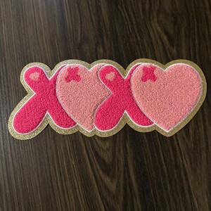 May include: A decorative patch featuring the letters "XOXO" in a heart-shaped design. The letters are in a vibrant pink color, with a gold glitter outline and a white border. The patch is likely for clothing or accessories.