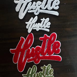 May include: A collection of six chenille patches featuring the word Hustle in various sizes and colors. There are two large white patches, two large red patches, and two smaller patches in white and olive green, all with a textured, fuzzy surface.