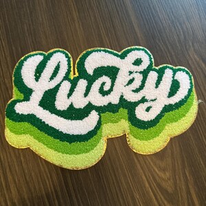 May include: A rug with the word "Lucky" in white, outlined in green and yellow. The rug has a layered design with shades of green and a gold border. The rug is placed on a wooden surface.