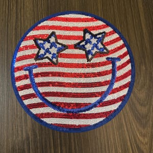 May include: A round, sequined patch with a patriotic design. The patch features red and white horizontal stripes, a blue border, two blue and white star-shaped eyes, and a blue smiling mouth. The design resembles a smiley face.
