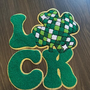 May include: A green and gold patch spelling "LUCK" with a shamrock design. The letters are green with gold trim, and the shamrock is green and white checkered. The patch is likely for St. Patrick's Day.