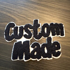 May include: A white felt patch with the words "custom made" in black, fluffy, block letters. The patch is on a dark wood-grain surface. The letters have a textured, fuzzy appearance.