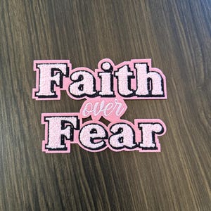 May include: Two pink patches with the words "Faith over Fear" in a bold, block letter style. The letters are outlined in black and have a fuzzy texture. The word "over" is in a smaller, cursive font.
