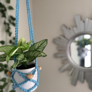 May include: A small white ceramic pot with a green plant, suspended by a light blue macrame hanger. The hanger features wooden beads and is hanging in front of a wall with a decorative mirror.
