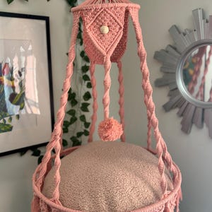 May include: A pink macrame hanging pet bed with a soft, beige cushion. The bed features intricate knotwork, tassels, and a decorative pom-pom. The design includes a wooden bead and is suspended from a ceiling hook.