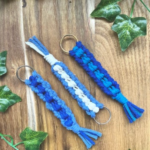 May include: Three blue and white macrame keychains with metal rings. The keychains are made with a braided cord and have a tassel at the end.