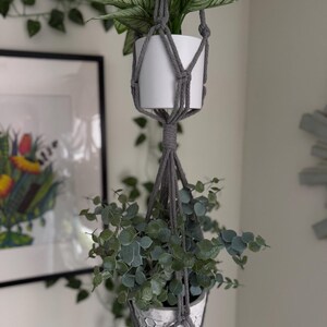 May include: A double-tiered macrame plant hanger in gray, holding two potted plants. The top pot is white, and the bottom pot is white with a textured pattern. Trailing greenery and a framed artwork are in the background.