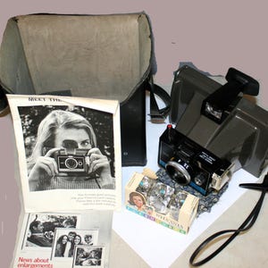 May include: Vintage Polaroid Land Camera with a black leather case, a flash attachment, and a box of Sylvania flashbulbs. The image also includes a brochure with the text "Meet the Polaroid Land Camera".