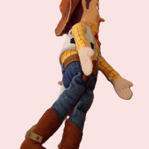 May include: A plush toy of a cowboy character, featuring a brown hat, yellow plaid shirt, denim jeans, and brown boots. The toy has a white vest with black detailing and tan gloves. The figure is posed in a walking stance.
