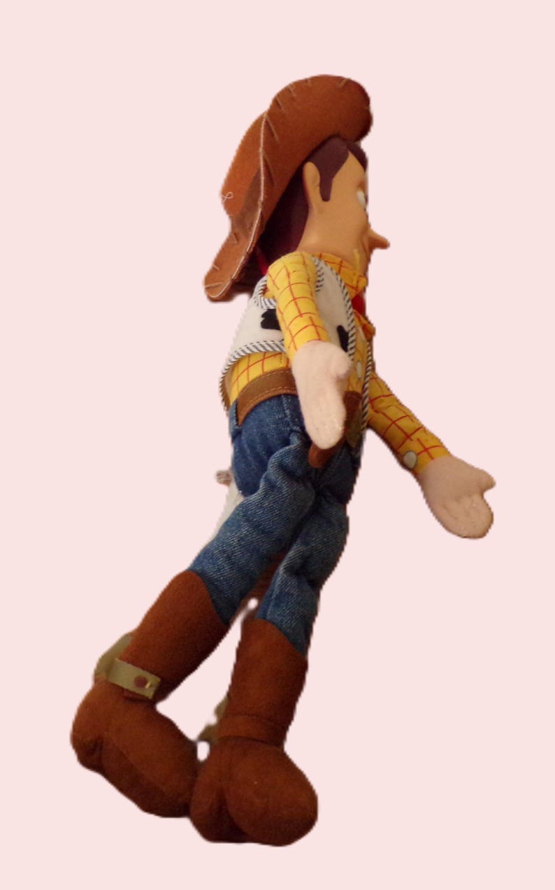 May include: A plush toy of a cowboy character, featuring a brown hat, yellow plaid shirt, denim jeans, and brown boots. The toy has a white vest with black detailing and tan gloves. The figure is posed in a walking stance.
