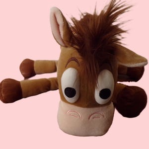 May include: A plush toy depicting a cartoon horse with brown fur, a light pink muzzle, and large, expressive eyes. The toy has a brown mane and four outstretched limbs, creating a playful and inviting appearance.