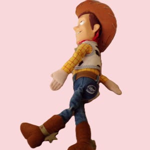 May include: A plush toy of the character Woody from the movie Toy Story. Woody is wearing a brown cowboy hat, a yellow plaid shirt, a vest with black spots, blue jeans, and brown boots with spurs. The toy is set against a light pink background.
