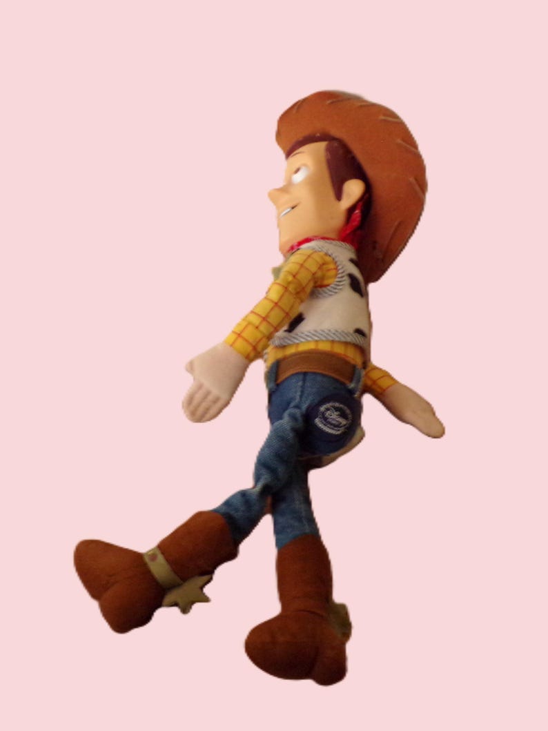 May include: A plush toy of the character Woody from the movie Toy Story. Woody is wearing a brown cowboy hat, a yellow plaid shirt, a vest with black spots, blue jeans, and brown boots with spurs. The toy is set against a light pink background.