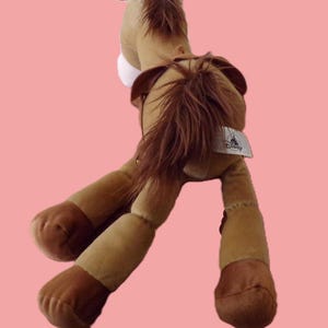 May include: A plush toy horse in shades of brown, tan, and dark brown, with a long mane and tail. The toy has a white patch on its chest and a Disney tag. The horse is posed with its legs extended.