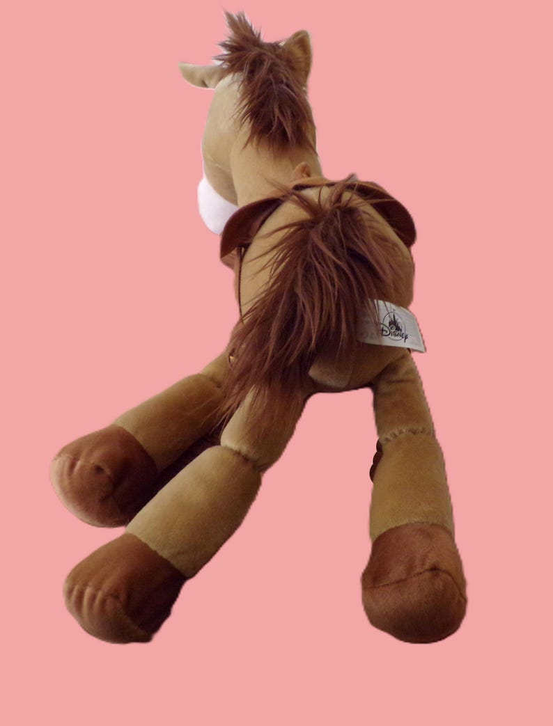 May include: A plush toy horse in shades of brown, tan, and dark brown, with a long mane and tail. The toy has a white patch on its chest and a Disney tag. The horse is posed with its legs extended.