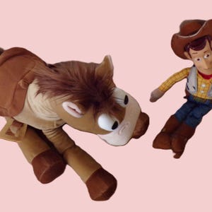 May include: A plush toy set featuring a brown horse and a Woody doll. The horse has a saddle and a brown mane. Woody is wearing a cowboy hat, vest, and boots. The background is a light pink color.