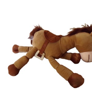May include: A plush toy horse in shades of brown, with a saddle and bridle. The toy has a light brown body, darker brown legs and mane, and a tan muzzle. The horse is lying down, with its legs extended.
