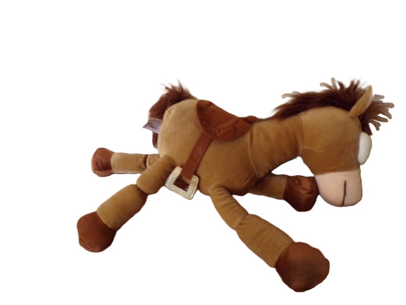 May include: A plush toy horse in shades of brown, with a saddle and bridle. The toy has a light brown body, darker brown legs and mane, and a tan muzzle. The horse is lying down, with its legs extended.