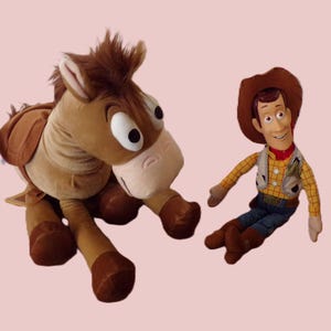 May include: A plush toy set featuring a brown horse and a Woody doll. The horse has a saddle and large eyes. Woody is wearing a cowboy hat, vest, and jeans. The toys are on a light pink background.