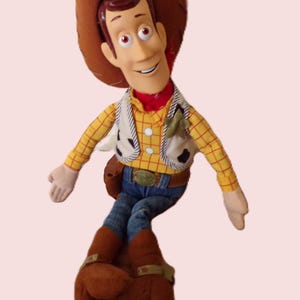 May include: A plush toy of a cowboy character, wearing a brown hat, yellow plaid shirt, denim jeans, and brown boots. The character has a red neckerchief and a white vest with black spots. The toy has a friendly expression.