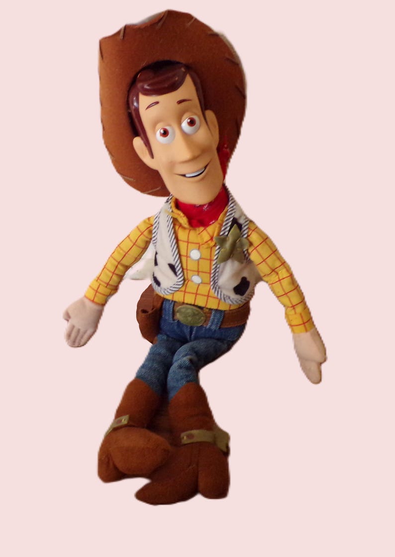 May include: A plush toy of a cowboy character, wearing a brown hat, yellow plaid shirt, denim jeans, and brown boots. The character has a red neckerchief and a white vest with black spots. The toy has a friendly expression.