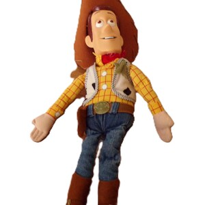 May include: A plush toy of a cowboy character, wearing a brown hat, yellow plaid shirt, denim jeans, and brown boots. The toy has a red neckerchief, a white vest, and a belt with a gold buckle. The toy's arms are outstretched.