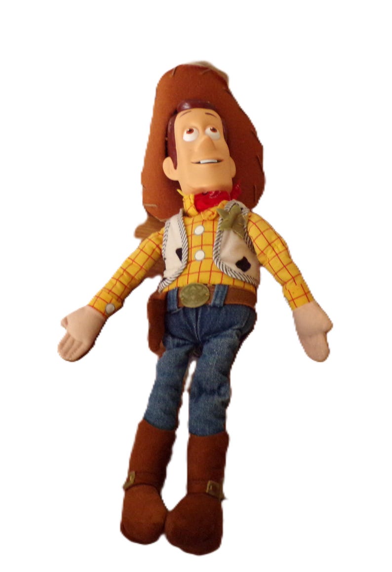 May include: A plush toy of a cowboy character, wearing a brown hat, yellow plaid shirt, denim jeans, and brown boots. The toy has a red neckerchief, a white vest, and a belt with a gold buckle. The toy's arms are outstretched.