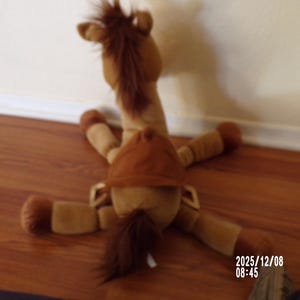 May include: A plush toy horse in shades of brown, lying on its back with legs spread. The toy has a brown mane and tail, and a brown saddle. The horse is positioned on a wooden floor.