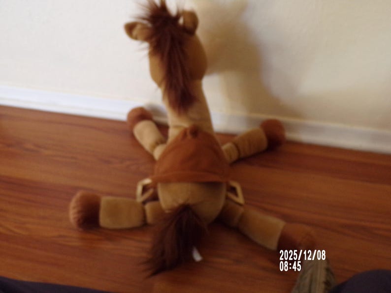 May include: A plush toy horse in shades of brown, lying on its back with legs spread. The toy has a brown mane and tail, and a brown saddle. The horse is positioned on a wooden floor.