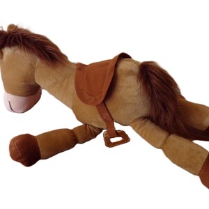 May include: A plush toy horse in a lying down position. The horse is primarily brown with a darker brown mane and tail. It has a saddle and bridle in a matching brown color. The horse's nose is pink.