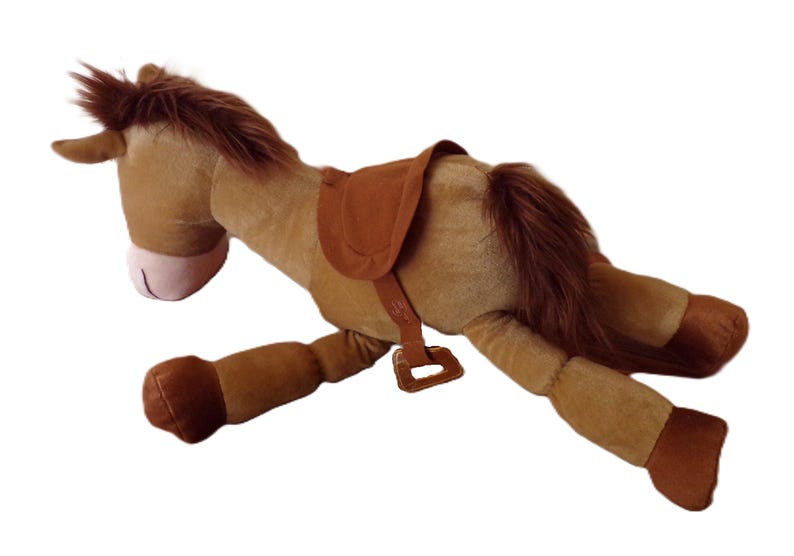 May include: A plush toy horse in a lying down position. The horse is primarily brown with a darker brown mane and tail. It has a saddle and bridle in a matching brown color. The horse's nose is pink.