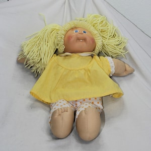 May include: A vintage doll with blonde yarn hair, blue eyes, and rosy cheeks. The doll wears a yellow dress with white polka dots and white lace trim. The doll's shorts are white with orange polka dots.