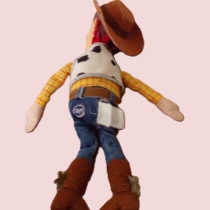 May include: A plush toy of a cowboy character, wearing a brown hat, a yellow plaid shirt, a cow-print vest, blue jeans, and brown boots. The toy has a white holster and a belt with a buckle. The cowboy doll is a popular children's toy.