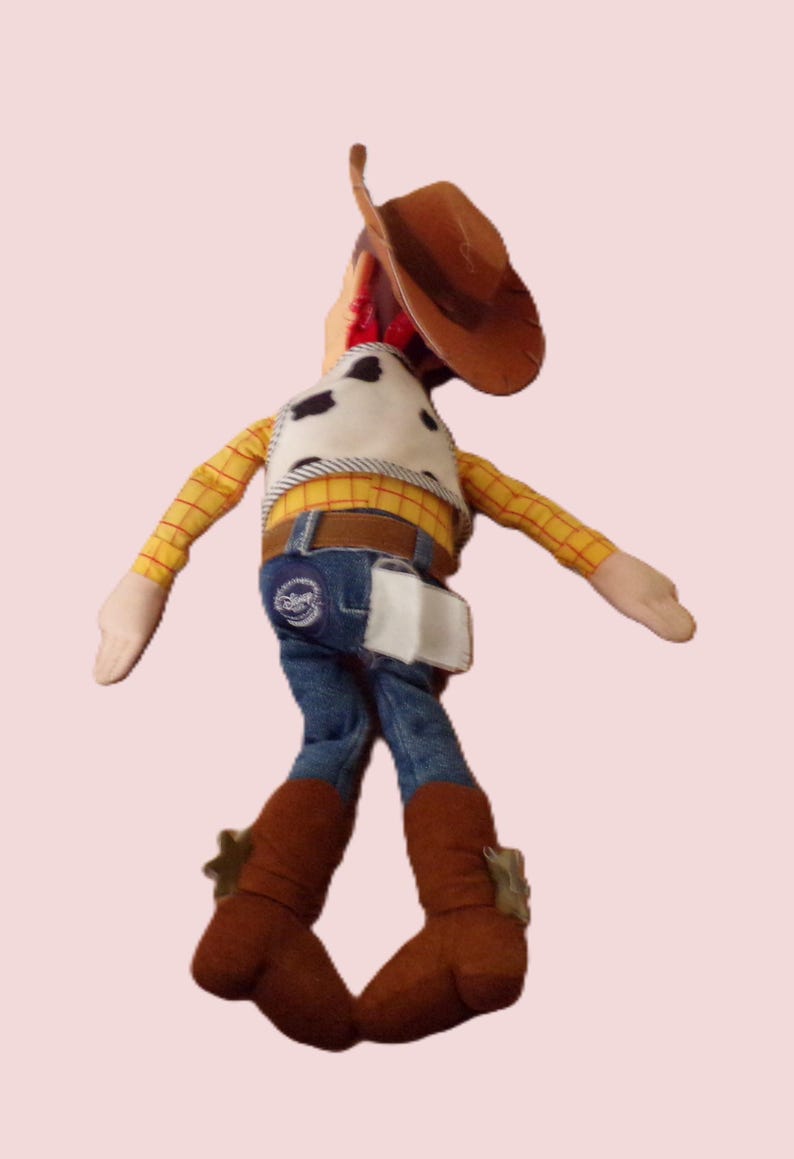 May include: A plush toy of a cowboy character, wearing a brown hat, a yellow plaid shirt, a cow-print vest, blue jeans, and brown boots. The toy has a white holster and a belt with a buckle. The cowboy doll is a popular children's toy.