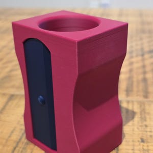 May include: A magenta pencil sharpener with a dark blue blade cover. The sharpener has a rectangular shape with curved sides and a circular opening at the top. It is on a wooden surface.