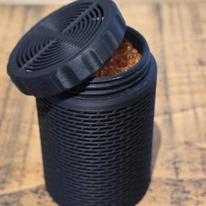 May include: A black, cylindrical container with a textured surface and a removable lid. The lid is partially open, revealing small, amber-coloured beads inside. The container is made of a matte black material.