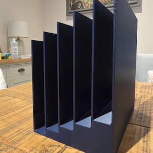May include: A dark blue desk tidy with five vertical compartments. The organiser is constructed from a solid material and is placed on a wooden table. The slots are designed to hold papers, files, or other office supplies.