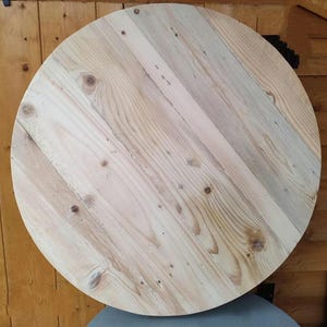 Op de afbeelding: Een ronde houten tafelblad gemaakt van gerecycled hout. Het hout heeft een natuurlijke lichtbruine kleur met wat donkerdere bruine knopen en nerven.